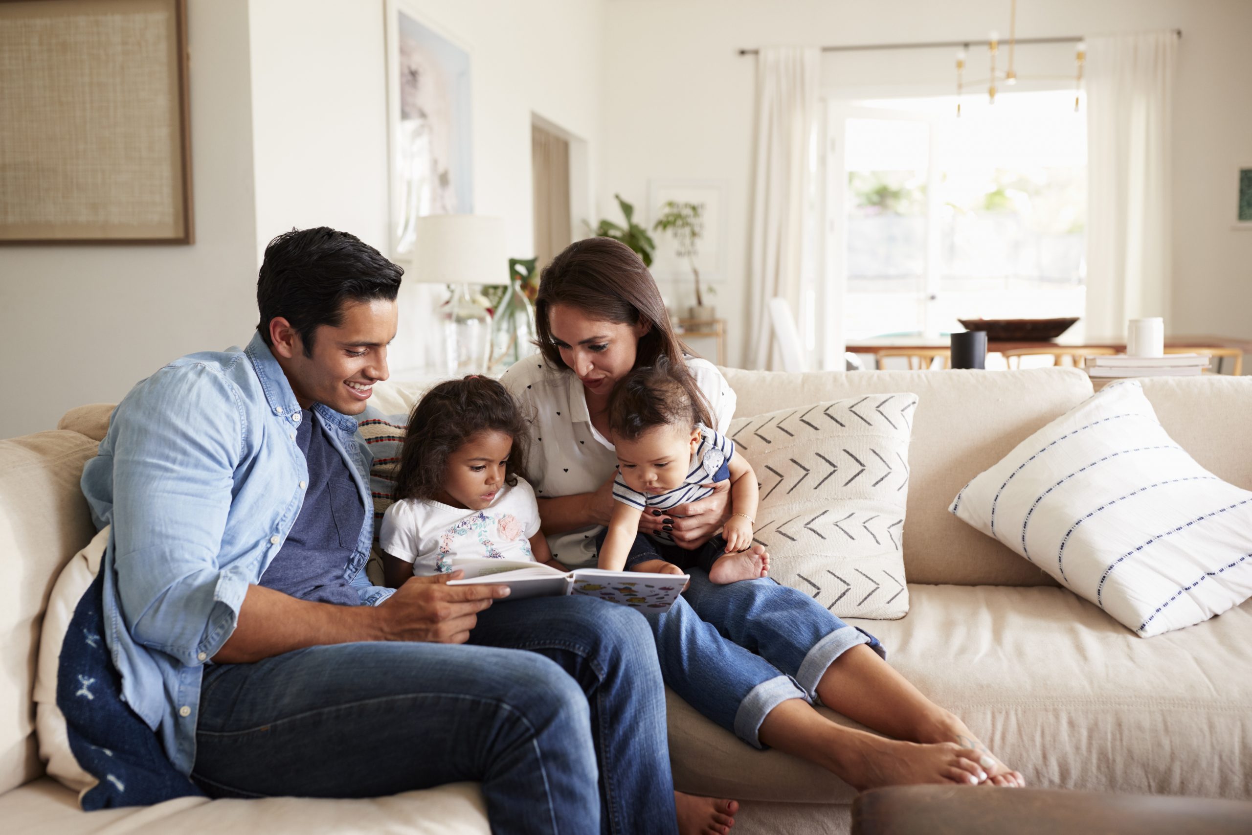 Hispanic couple sitting on the sofa reading a book at home with their baby son and young daughter family health history month