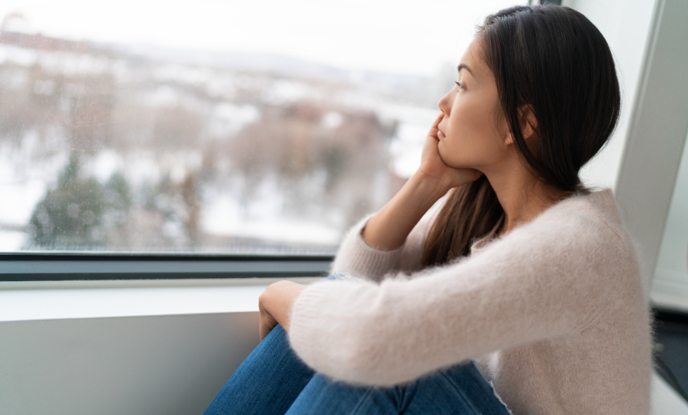 Asian woman with seasonal depression in pink sweater stares out window with snow on the ground outside